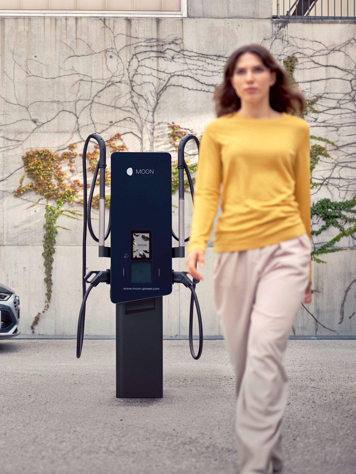 Frau geht vor einer Wallbox vorbei mit einem E-Auto im Hintergrund