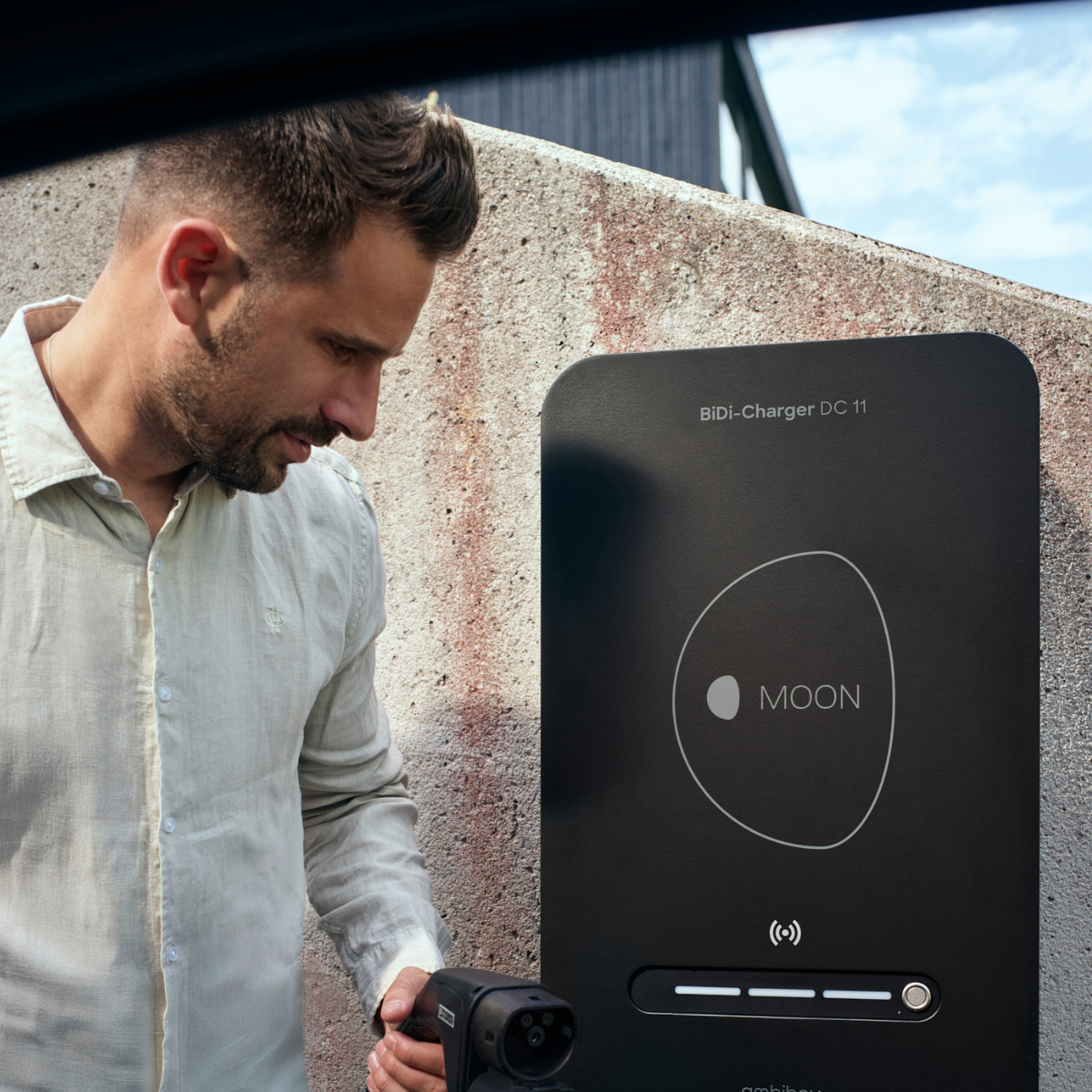 Mann mit bidirektionaler Wallbox MOON BiDi Charger vor Auto