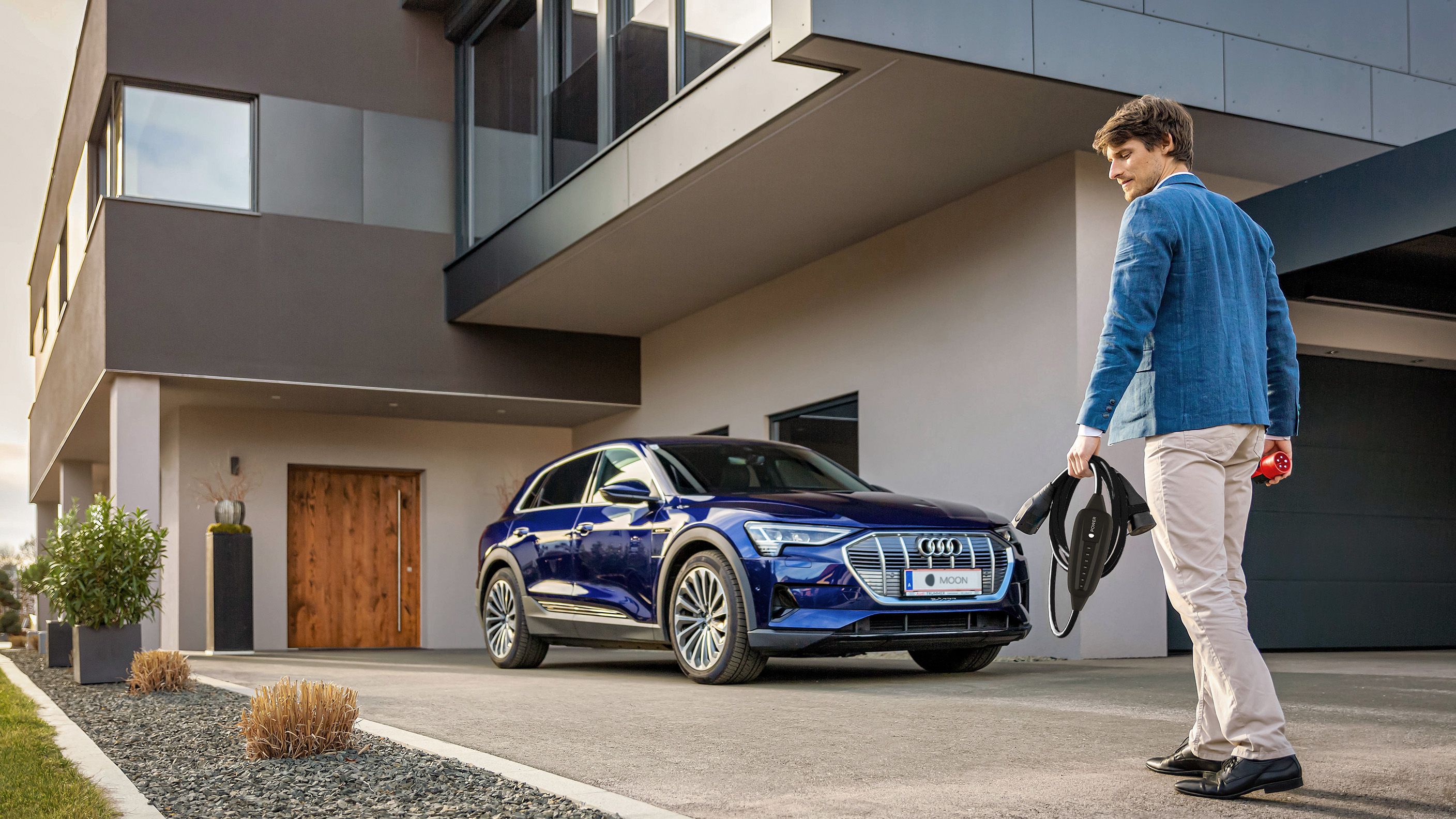 Mann geht mit einem POWER2Go Kabel in der Hand auf ein E-Auto bei einem Haus zu