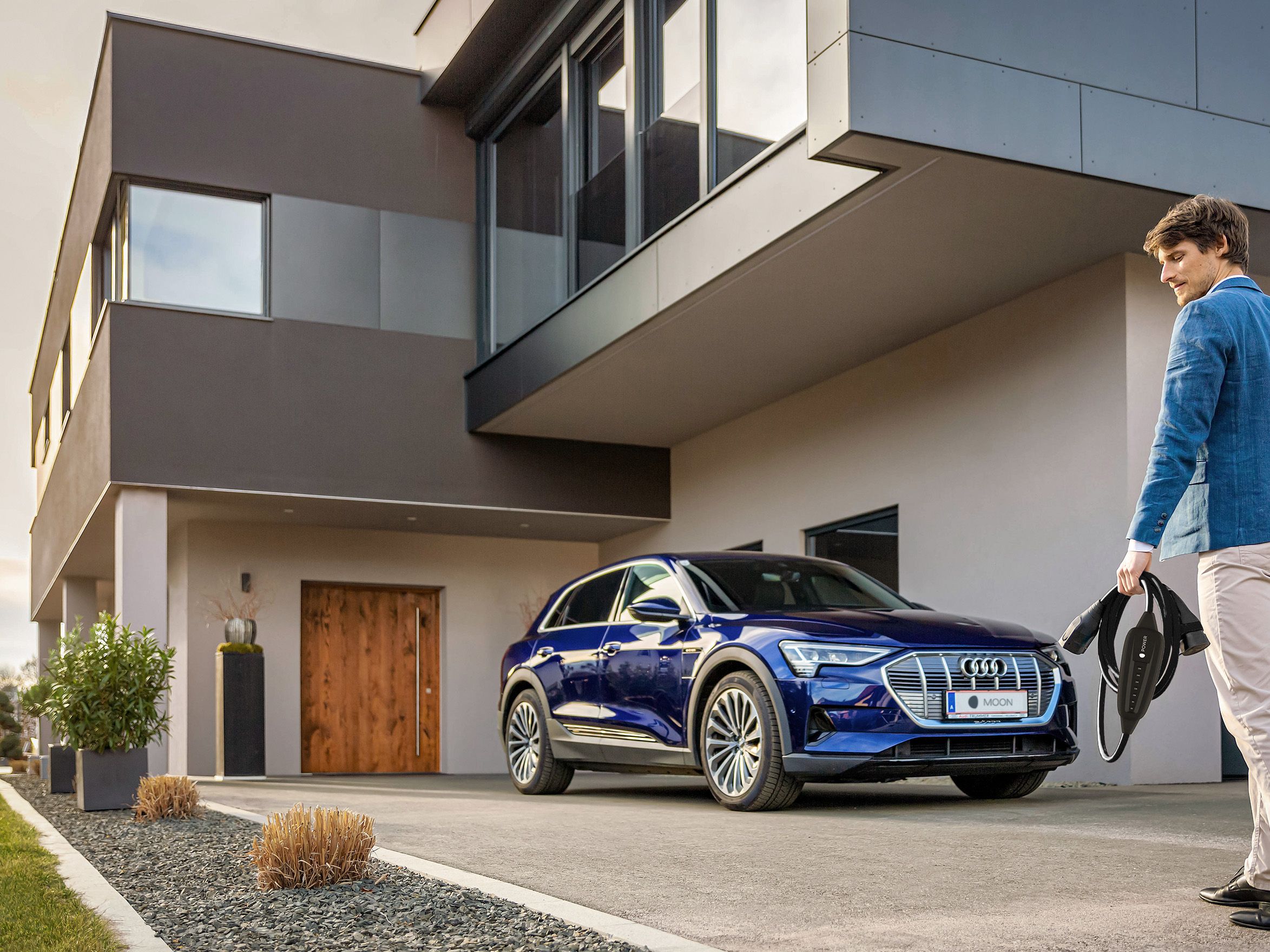 Mann geht mit einem POWER2Go Kabel in der Hand auf ein E-Auto bei einem Haus zu