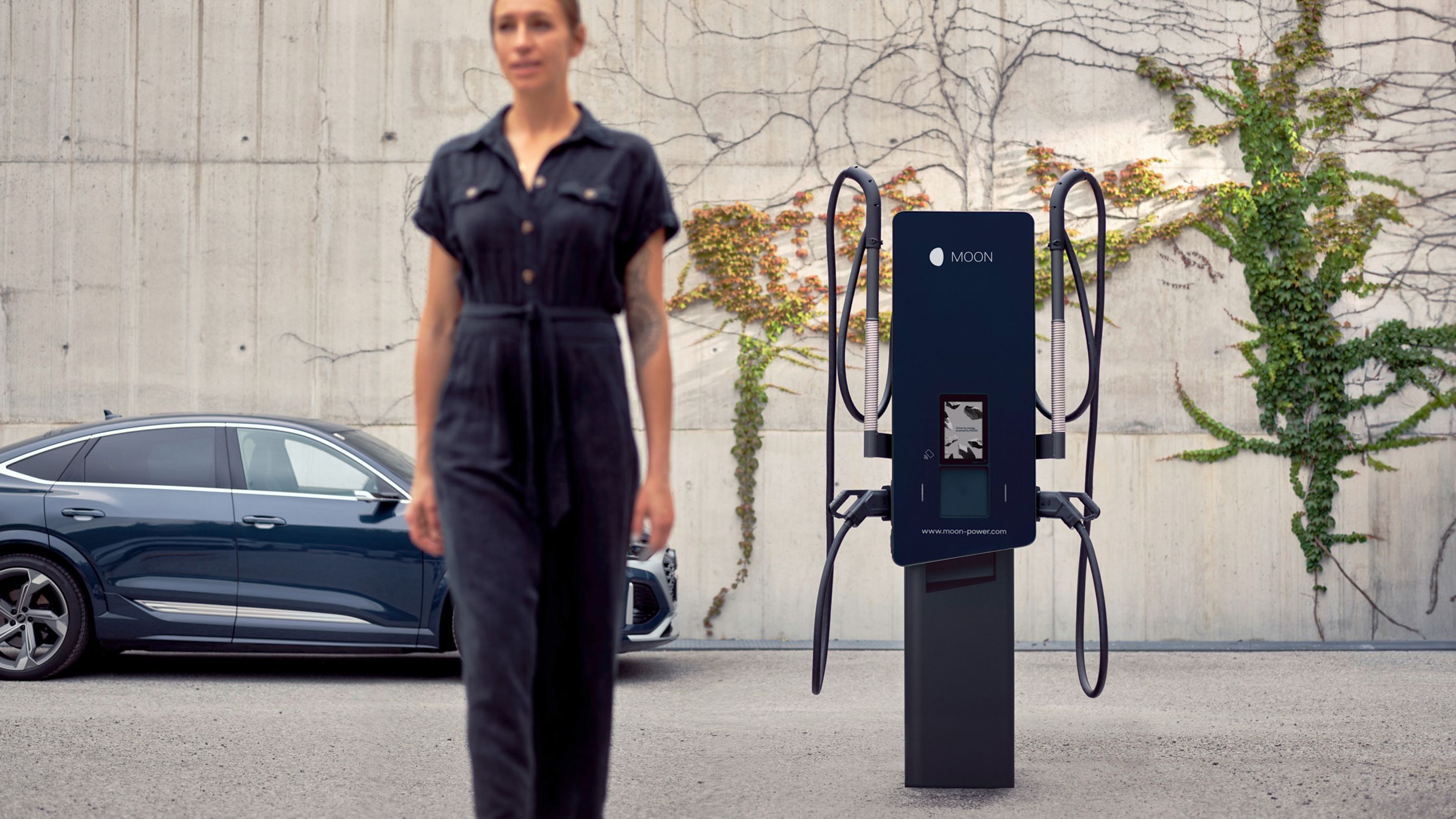 Frau geht vor einer Wallbox vorbei mit einem E-Auto im Hintergrund