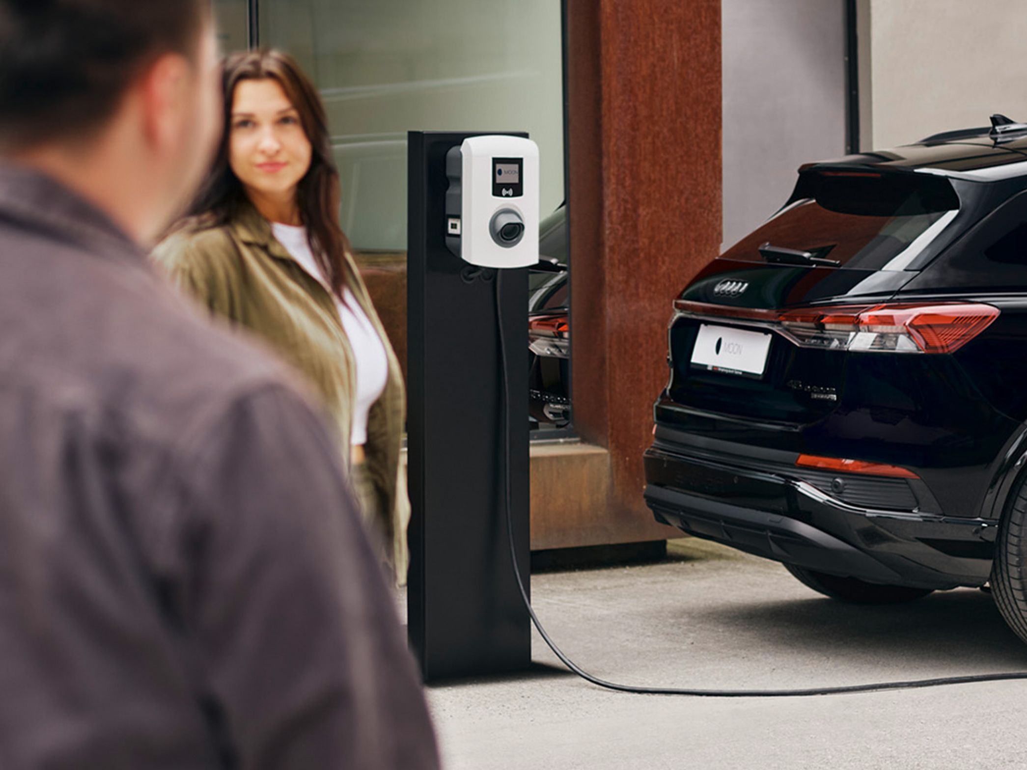 Ein E-Auto lädt an einer Wallbox mit zwei Menschen im Vordergrund