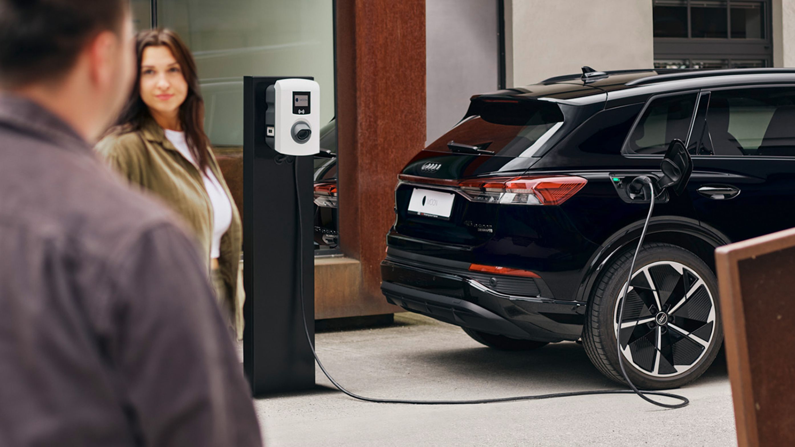 Ein E-Auto lädt an einer Wallbox mit zwei Menschen im Vordergrund